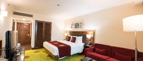 A hotel room with a bed, a red sofa, a desk with a laptop, a TV, and a green carpet.