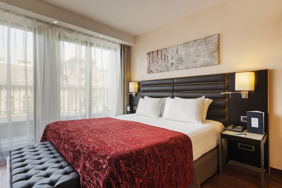 A hotel room with a large bed, a bedside table with a lamp, a bench, and a view of the outdoors through sheer curtains.