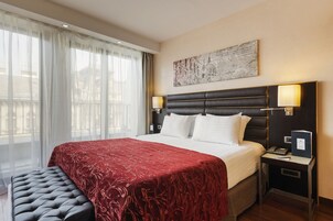 A hotel room with a large bed, a bedside table with a lamp, a bench, and a view of the outdoors through sheer curtains.