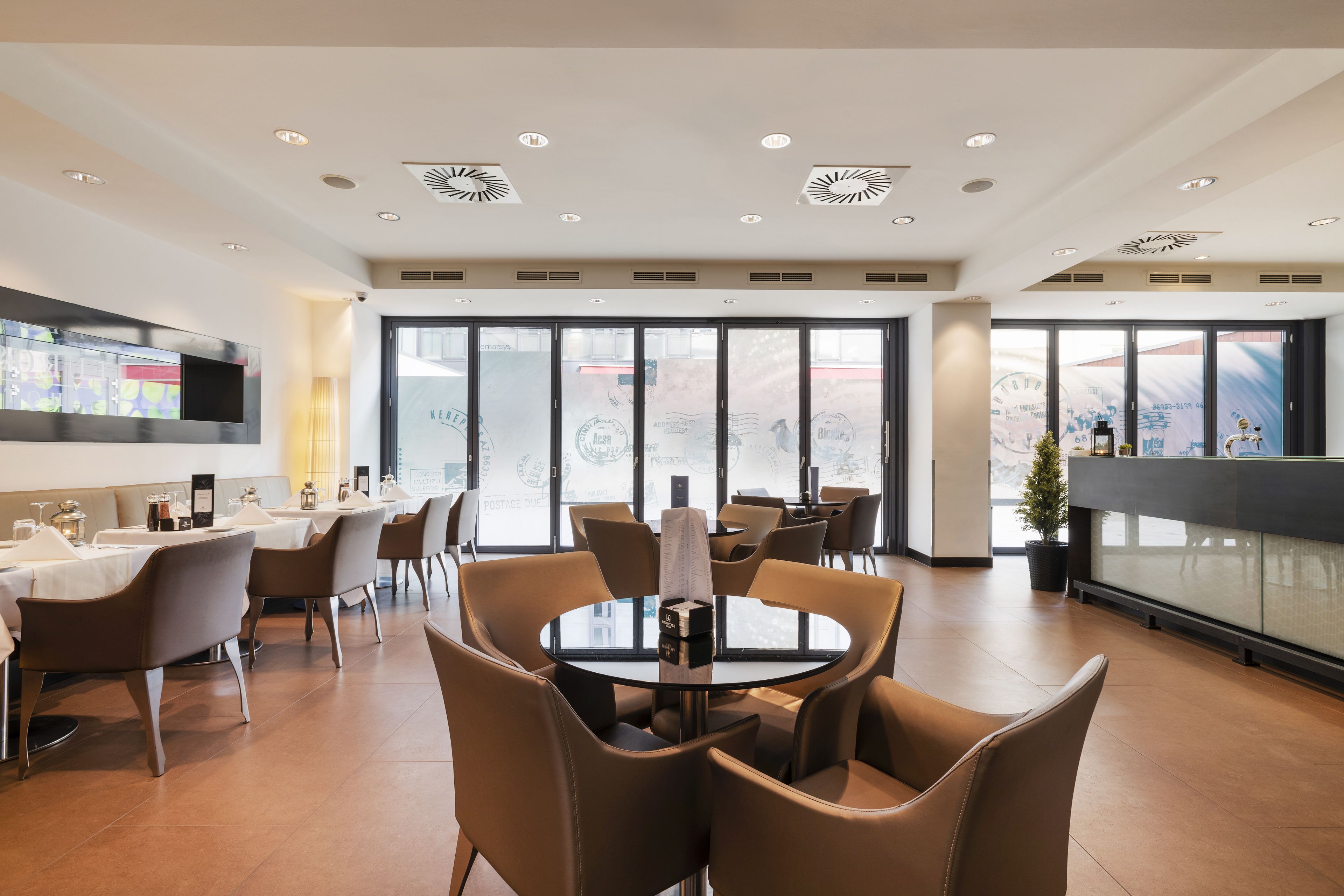 A modern dining area with round tables and brown chairs, large windows with a view, and a flat-screen TV mounted on the wall.