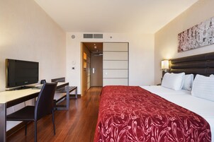 Quarto de hotel com cama, mesa de escritório, televisão, cadeira e mesa de cabeceira.