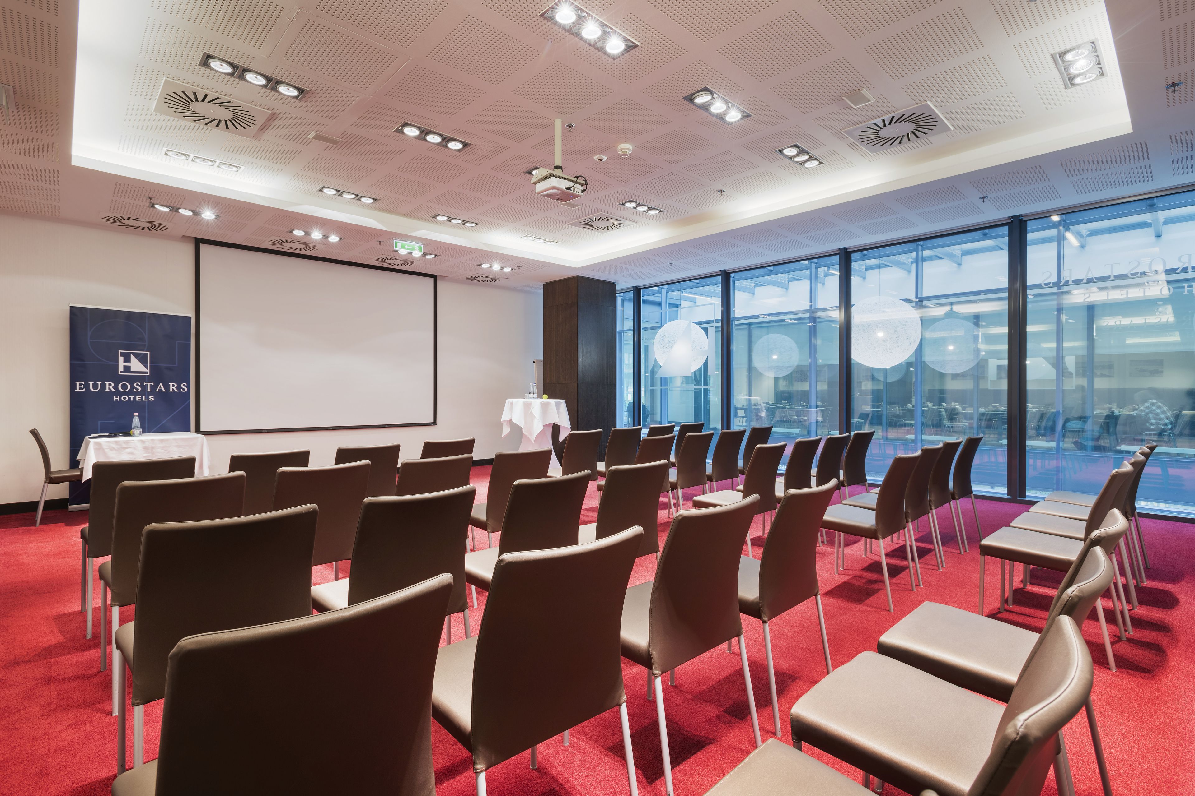 Un salón de conferencias con filas de sillas y mesas, una pantalla de proyector y un ventanal con vistas a la ciudad.