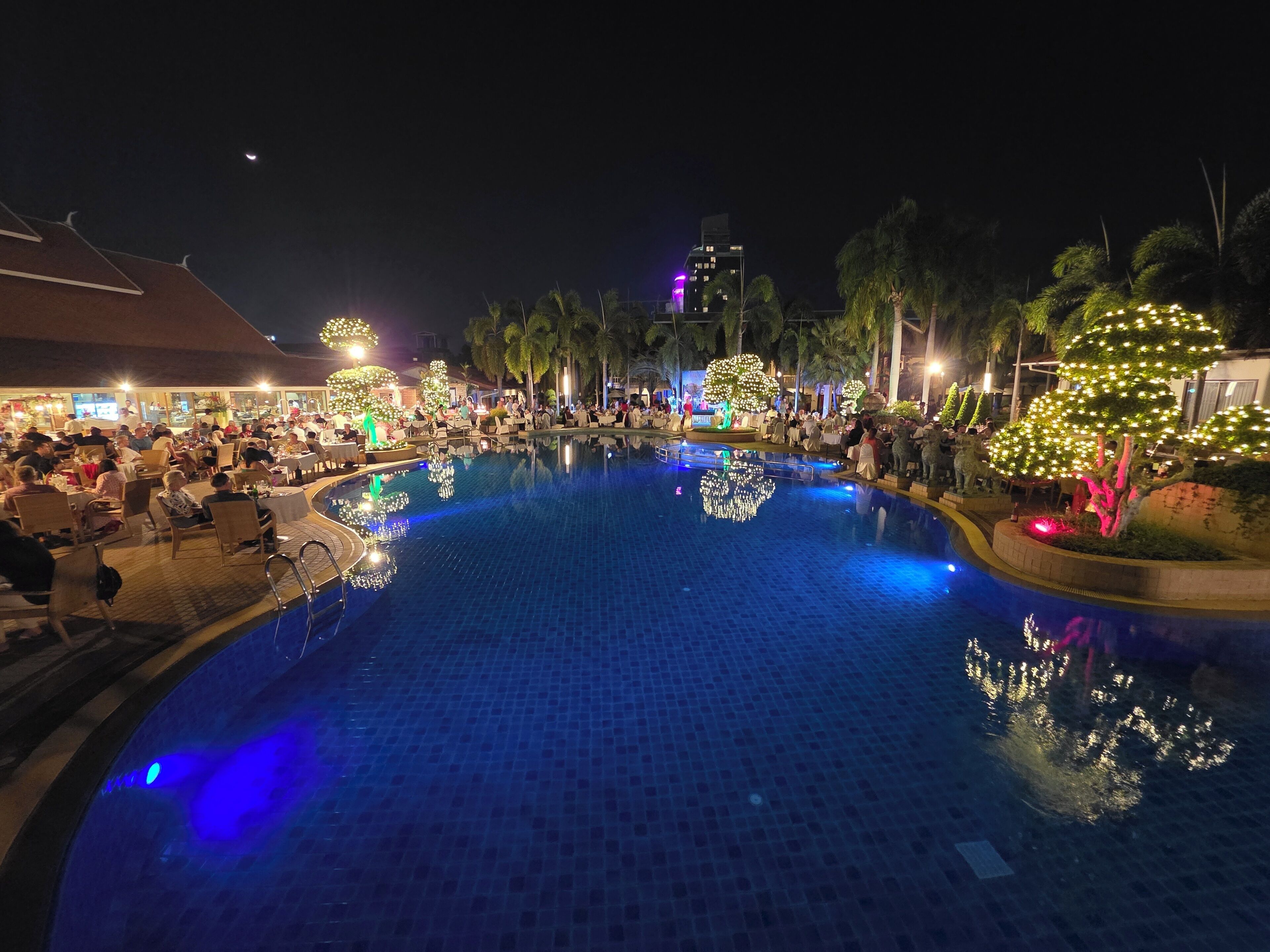 A nighttime pool area with tiled flooring, surrounded by palm trees and illuminated by decorative lights.