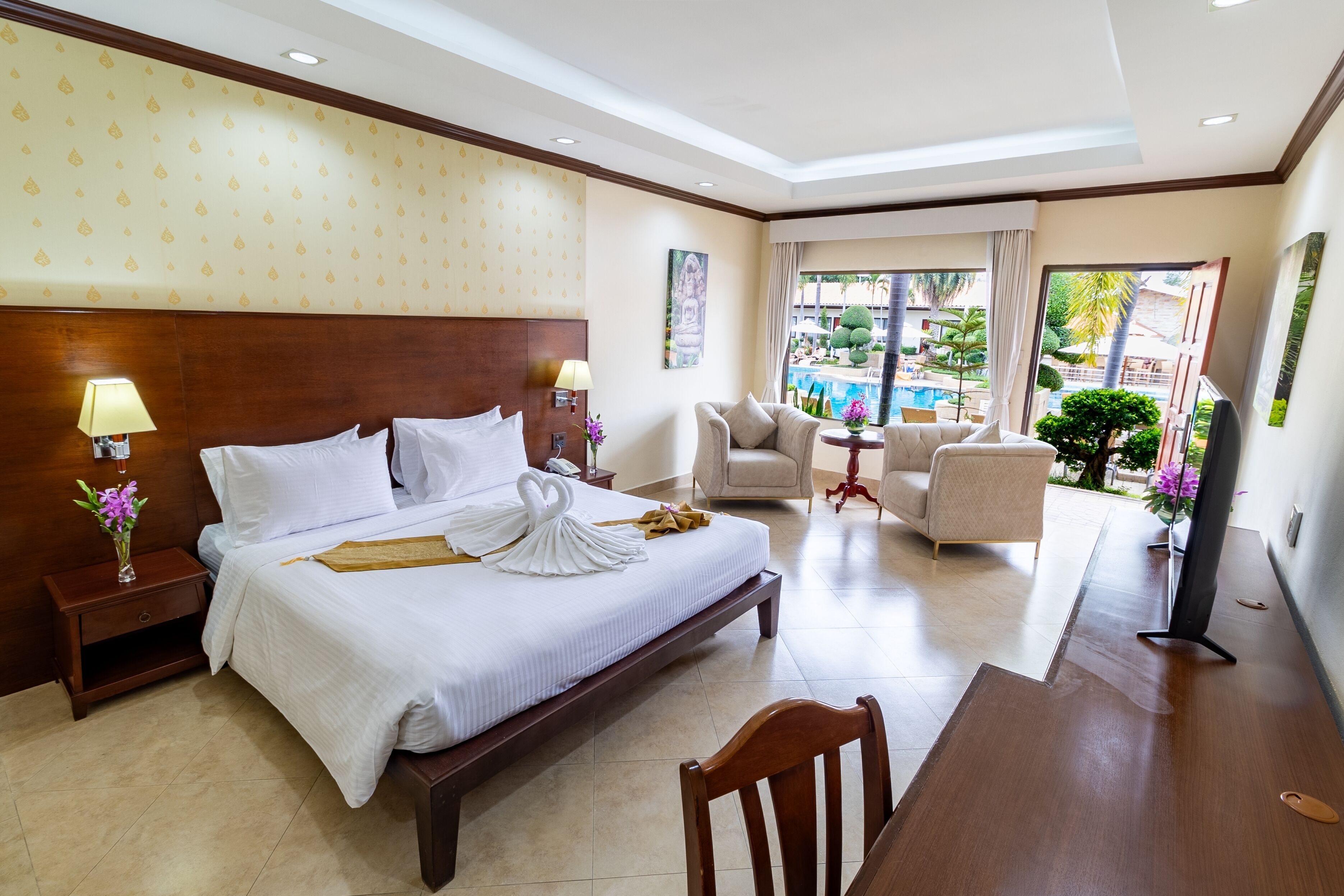 A hotel room with a large bed, a desk, a chair, a TV, and a view of a swimming pool and greenery.