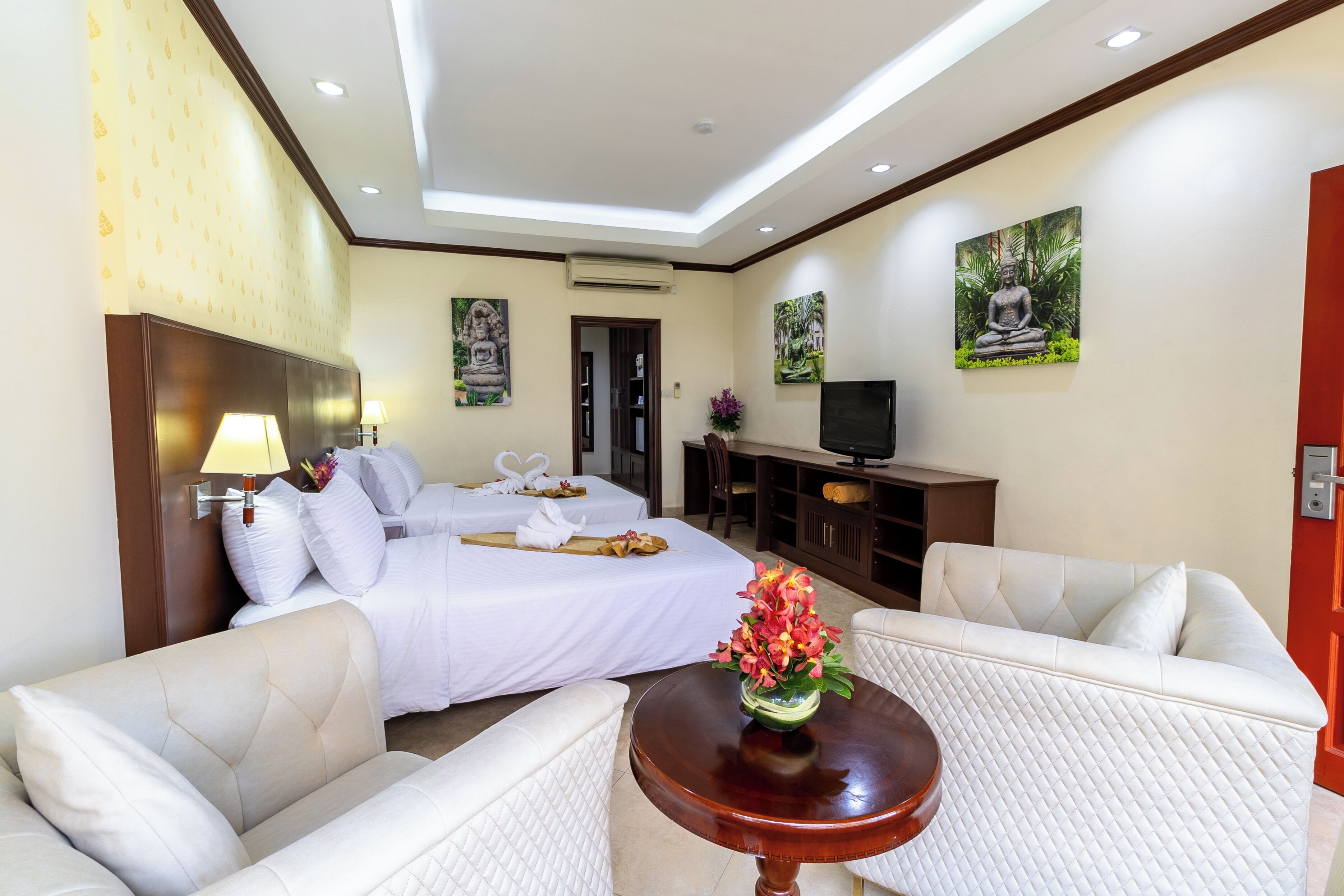 A hotel room with a bed, two white sofas, a small table with a vase, and a TV on a wooden cabinet.