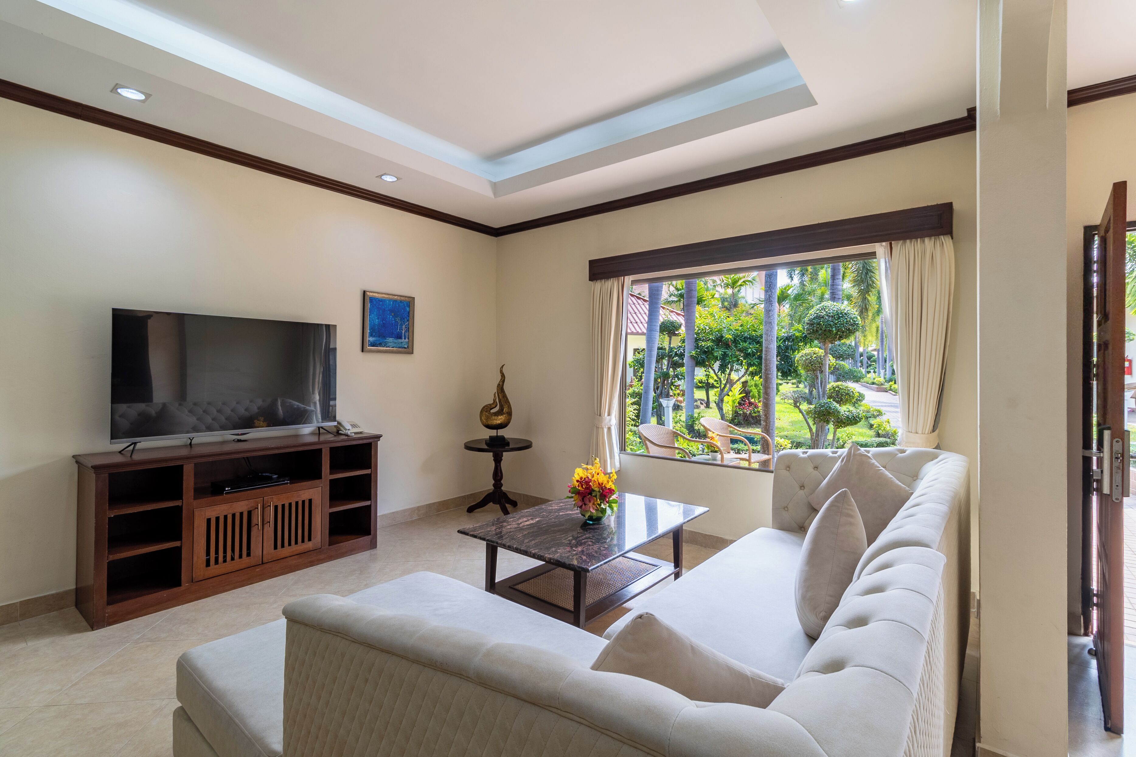A living room with a sofa, a coffee table, a TV on a wooden stand, and a window with a view of greenery.
