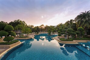 A large, clear swimming pool surrounded by well-manicured trees and palm trees, with a sunset in the background. - Thai Garden Resort (Pattaya)