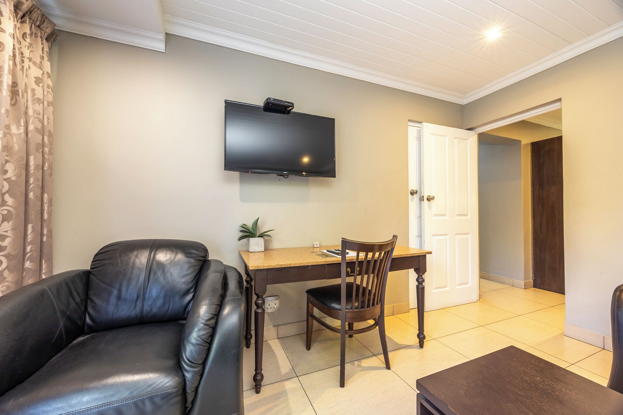 A living room with a leather sofa, a wooden dining table, a TV mounted on the wall, and a door leading to another room.