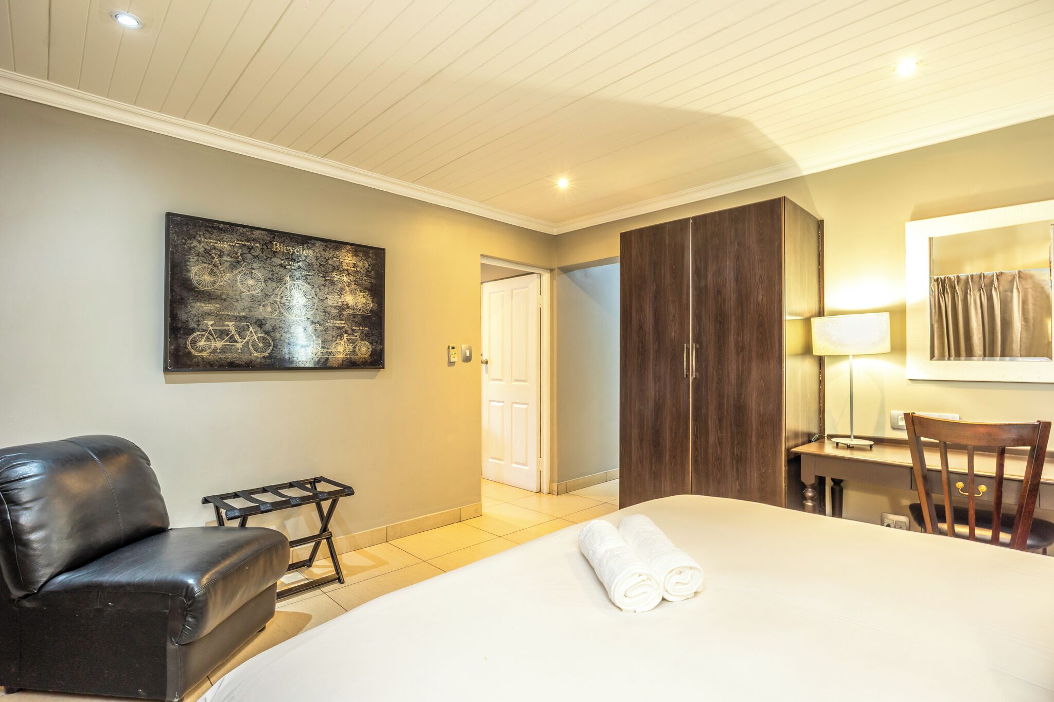 A hotel room with a bed, a black leather chair, a desk with a lamp, a wooden wardrobe, and a wall-mounted bicycle artwork.