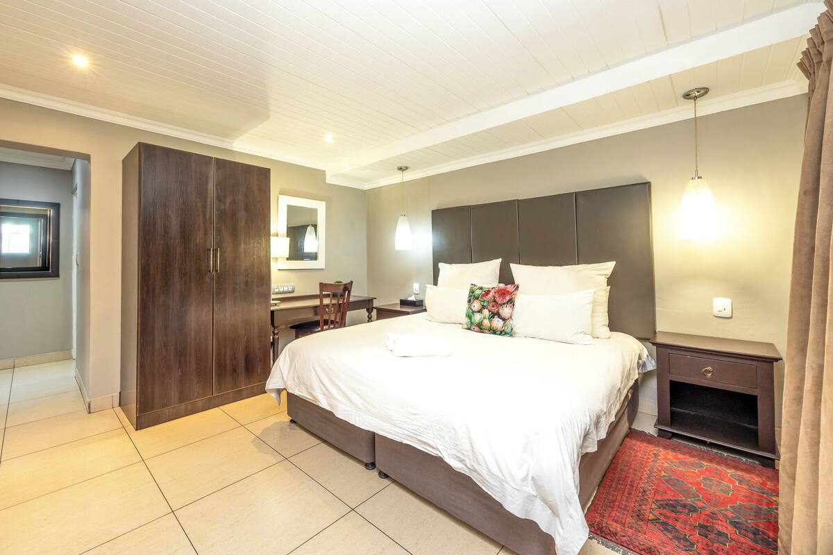 A hotel room with a large bed, a desk, a wardrobe, and a red patterned rug.