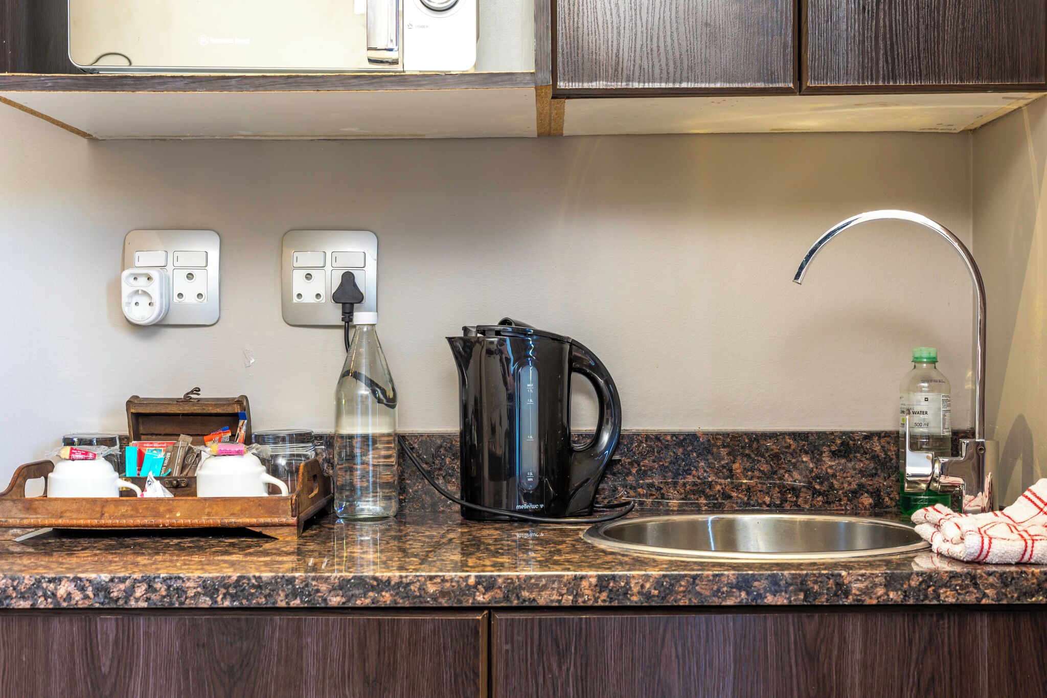 A kitchen with a granite countertop, a coffee maker, a kettle, a sink, and a bottle of water.
