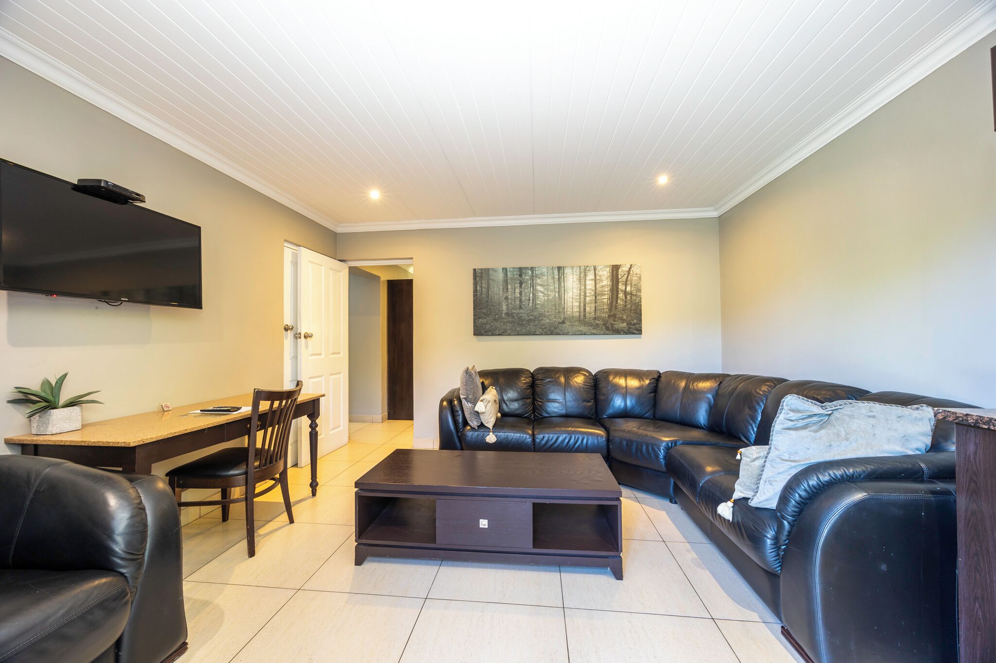 A living room with a sofa, a coffee table, a TV mounted on the wall, a dining table with chairs, and a painting on the wall.