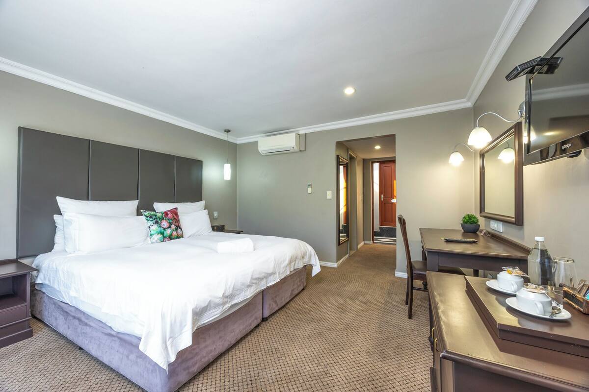 A hotel room with a large bed, a desk, a mirror, and a small kitchenette area.