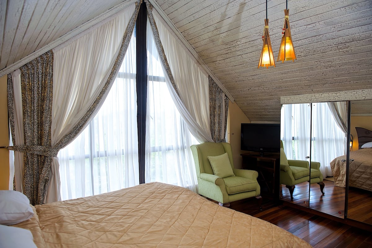 A bedroom with a large bed, two green armchairs, a TV, and a mirror reflecting the room.