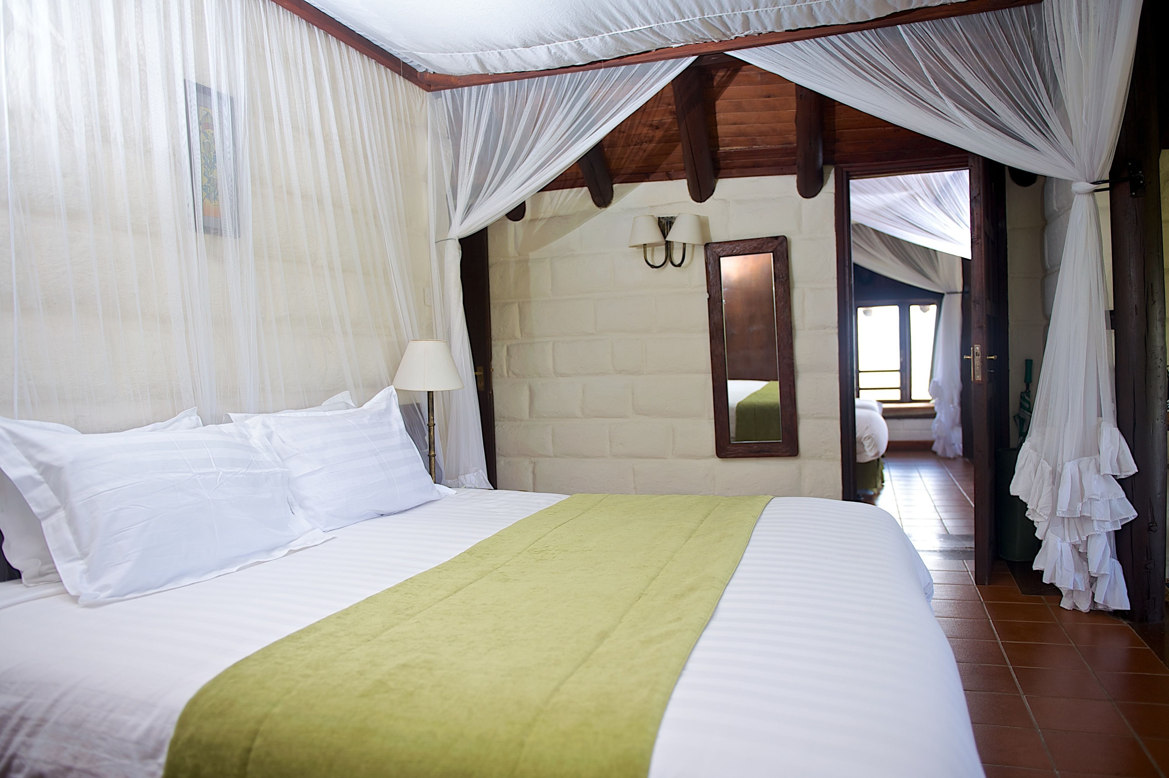 Une chambre d’hôtel avec un lit, des rideaux blancs, un plaid vert, un plafond en bois et un miroir au mur.