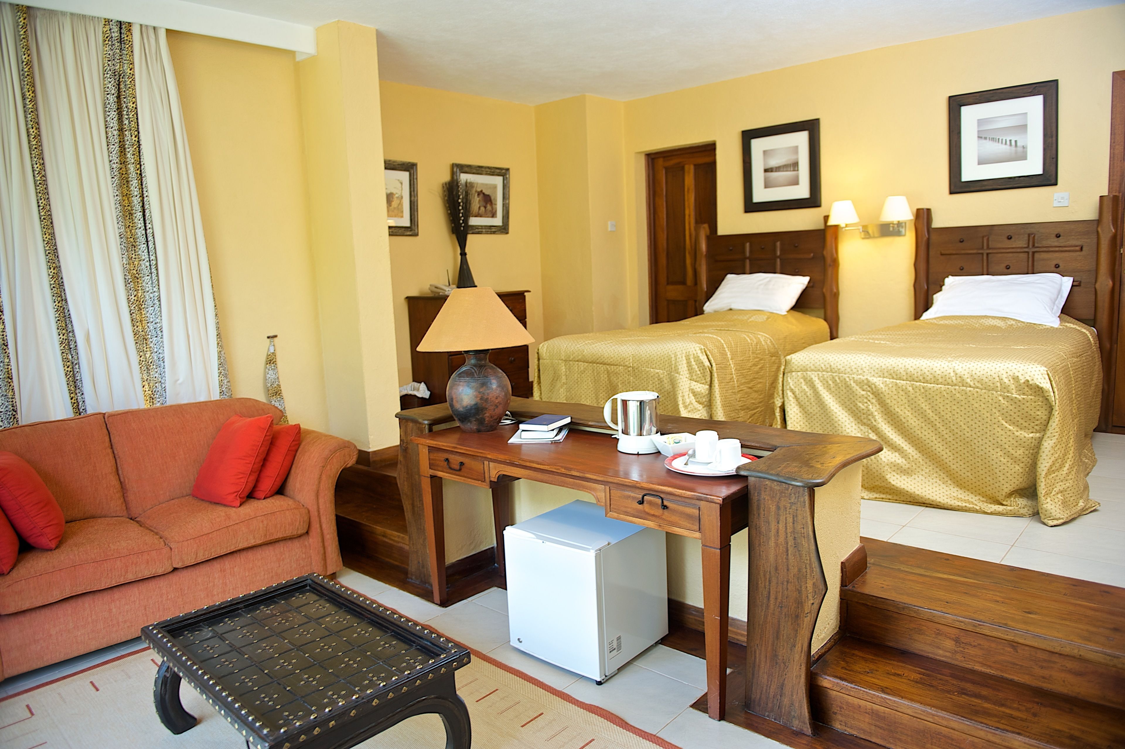 Habitación de hotel con sofá, escritorio con lámpara, cafetera, dos camas y un refrigerador pequeño.