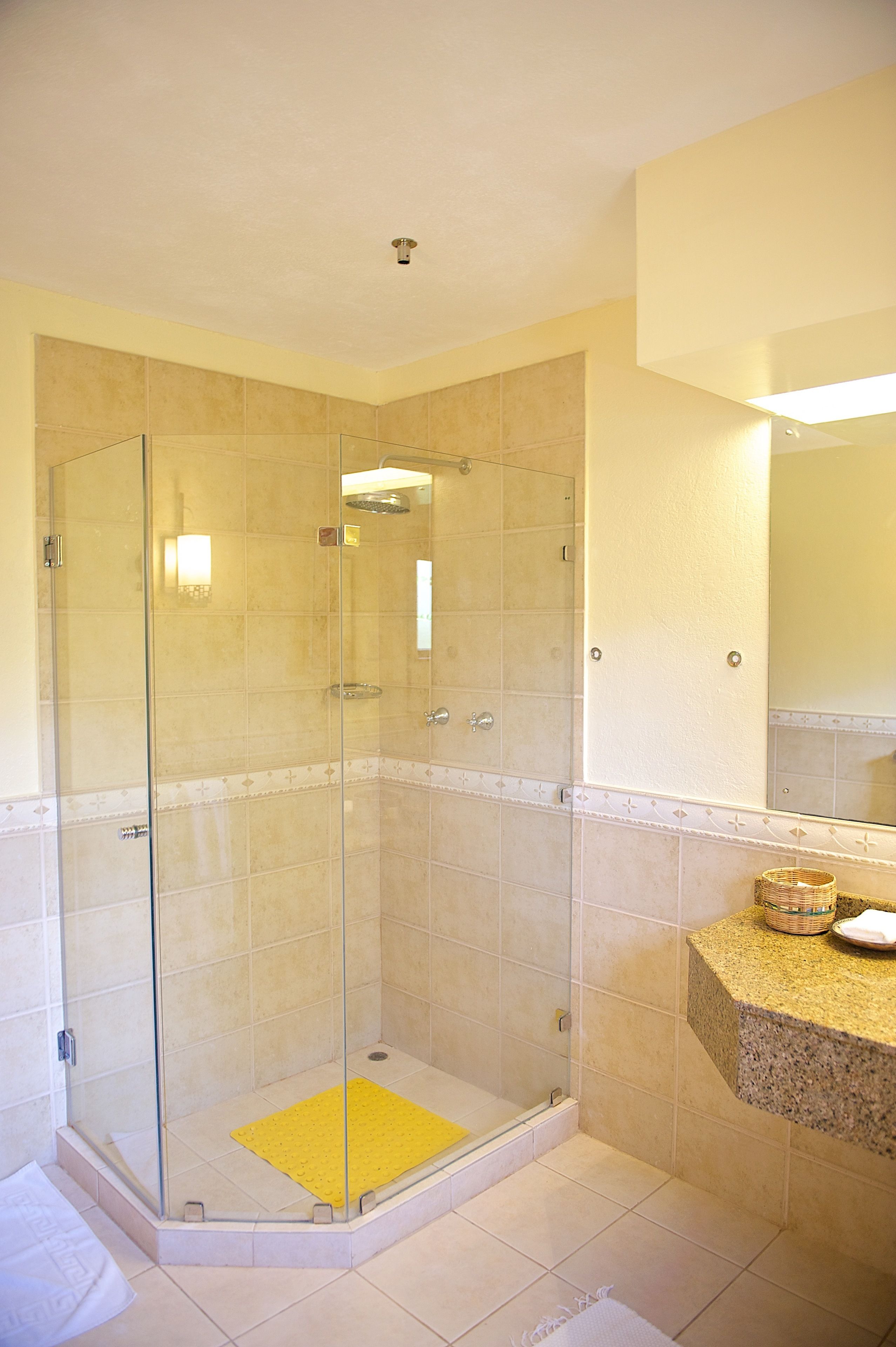 A glass-enclosed shower with a yellow mat, a granite countertop, and a mirror above it.