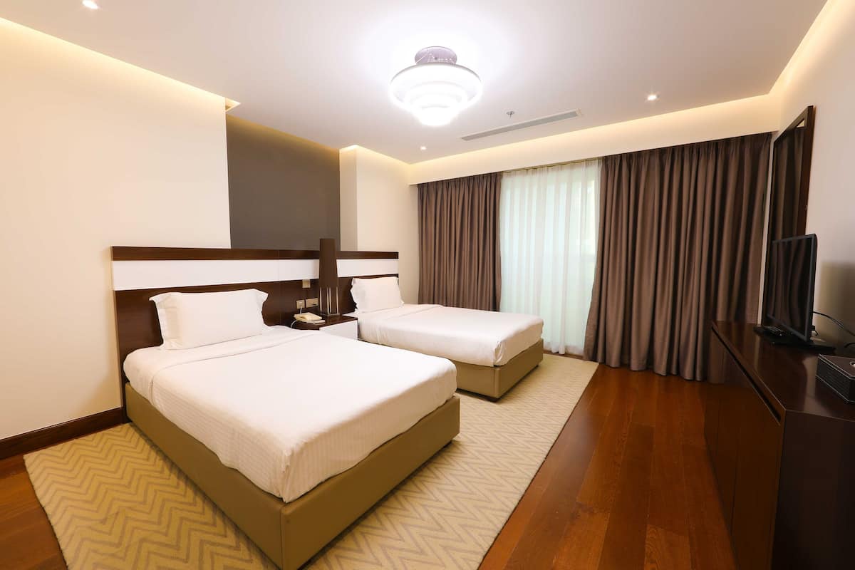 A hotel room with two beds, a flat-screen TV, a desk, and a large window with curtains.