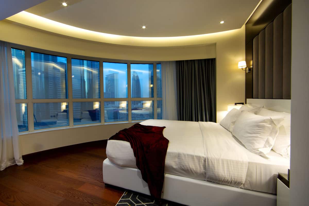 A modern hotel room with a curved ceiling, large windows offering city views, a white bed with a red throw, and a wooden floor.