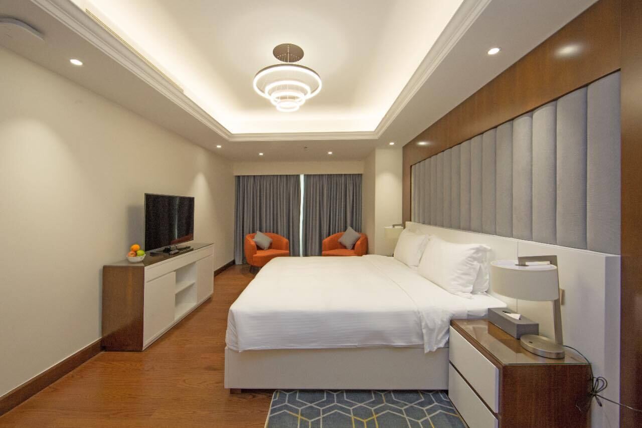 A hotel room with a large bed, a TV, two chairs, a desk, and a modern ceiling light.