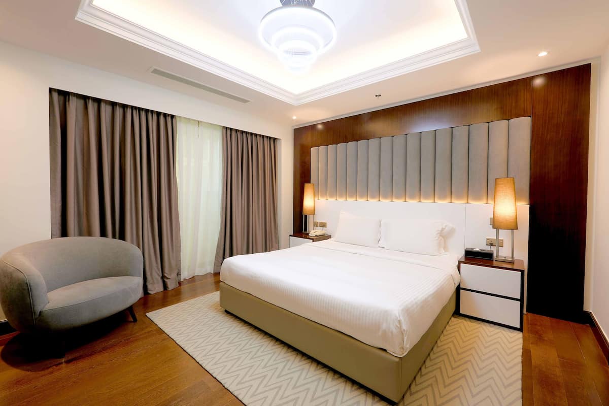 A modern hotel room with a large bed, a grey armchair, a wooden headboard, and a nightstand with lamps.