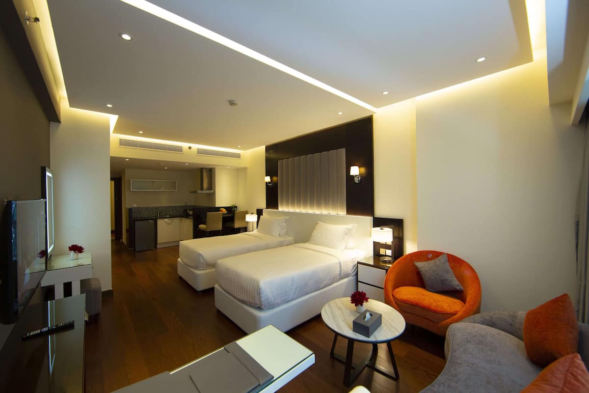 A modern hotel room with two beds, a sitting area with a sofa and armchair, a small table, and a TV.