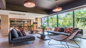 Una sala de estar moderna con sofá, sillón y mesa de centro, ventanas amplias con vistas a la vegetación, y una zona de cocina al fondo.