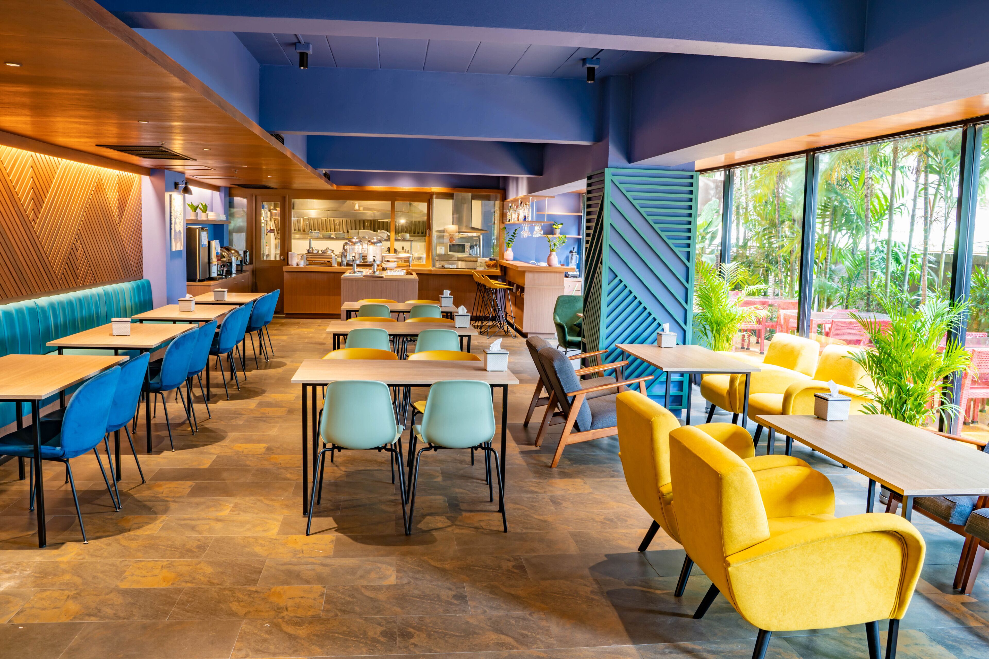 A modern restaurant interior with multiple dining tables and chairs, a kitchen area, and large windows offering a view of greenery.