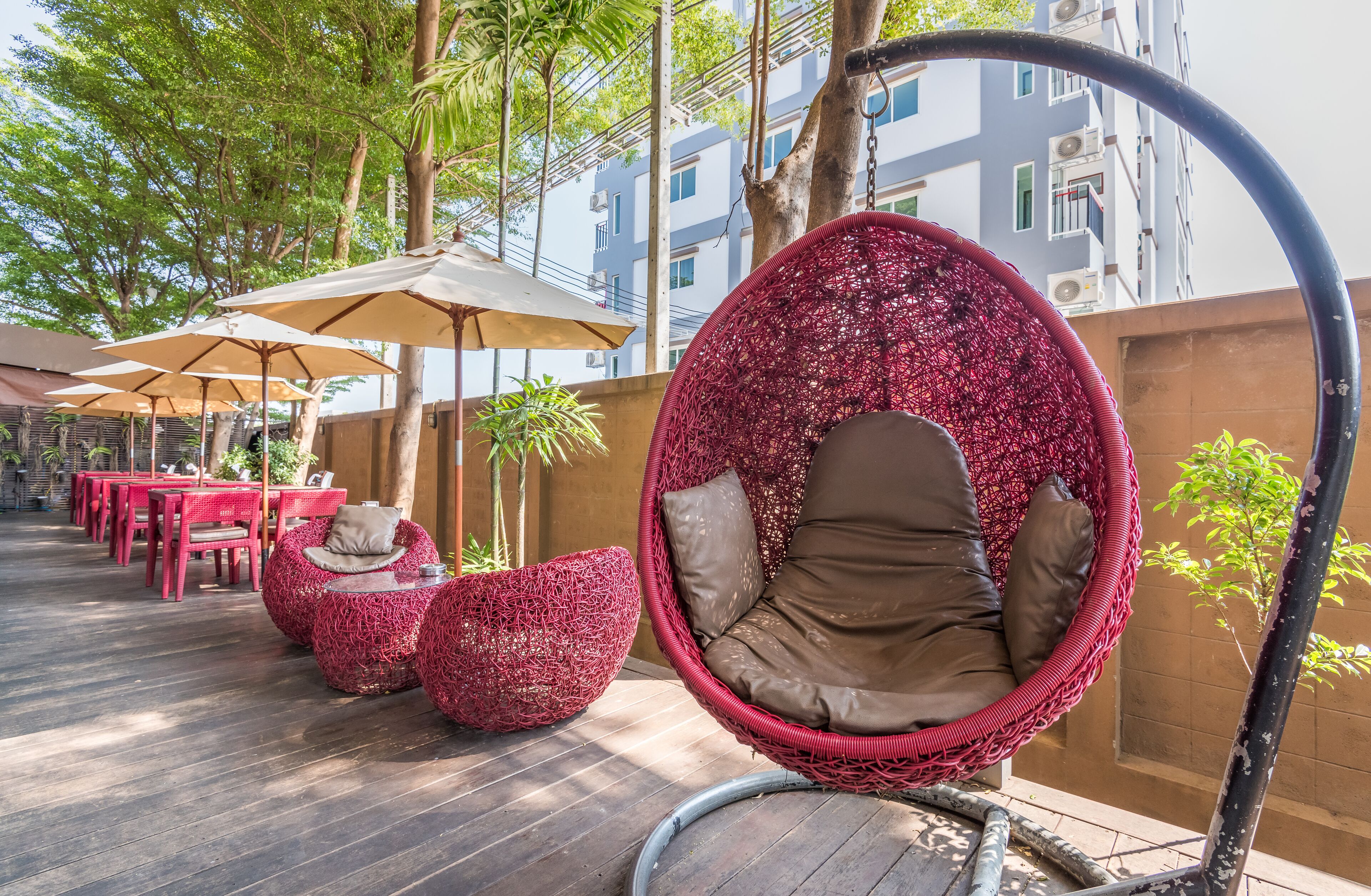 A seating area with red chairs and a hanging chair, surrounded by trees and a building in the background.