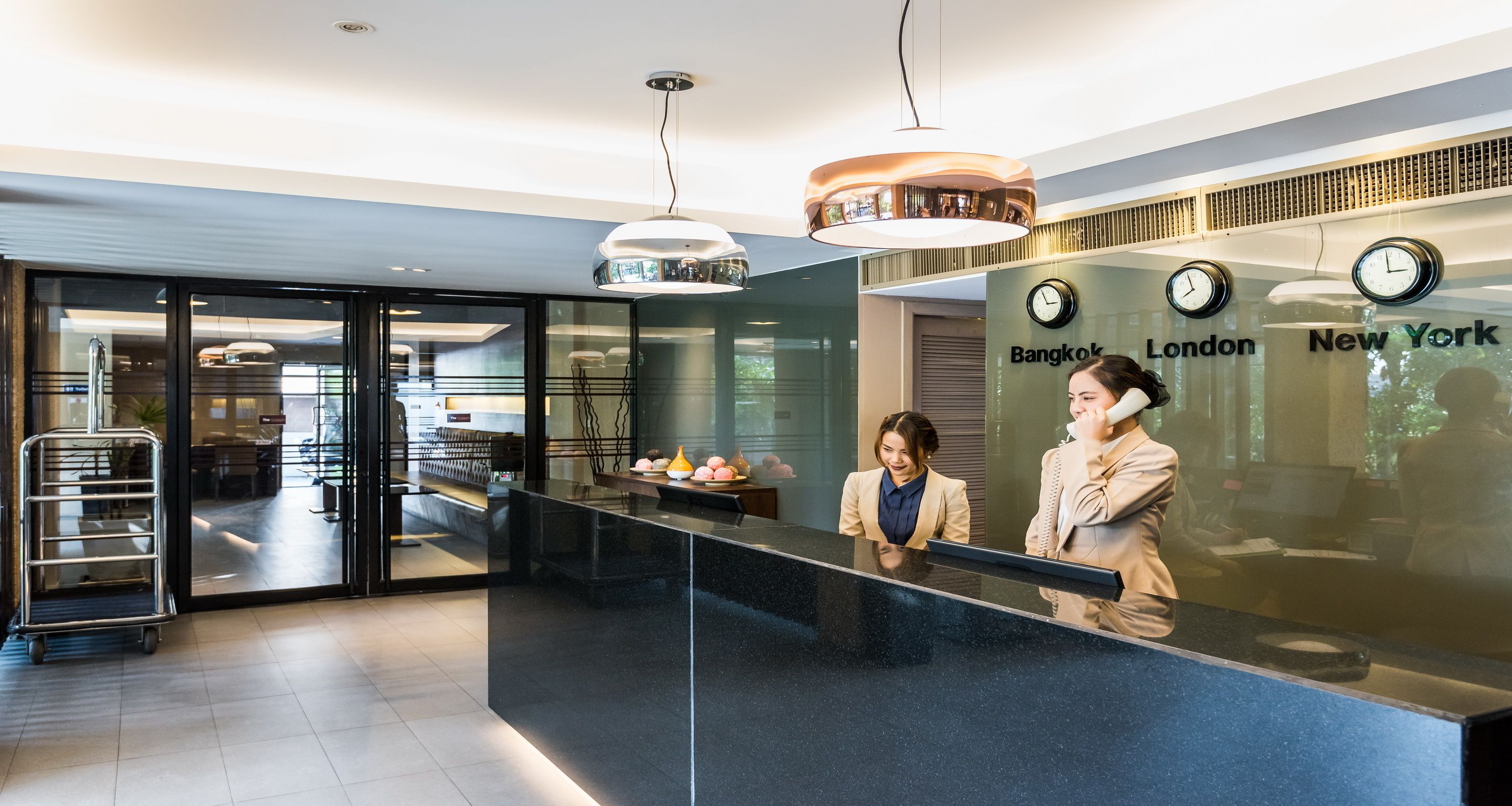 A reception desk with a glass partition, clocks displaying times for Bangkok, London, and New York, and a cart with a tray of items.