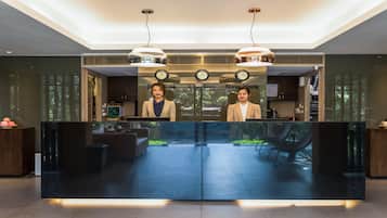Un mostrador de recepción con dos recepcionistas, un reloj digital y vistas al área de estar exterior.