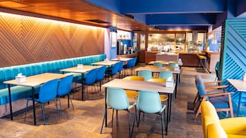 Een modern restaurant met blauwe en gele zitplaatsen, een houten accentmuur en een zichtbare keuken door een glazen wand.