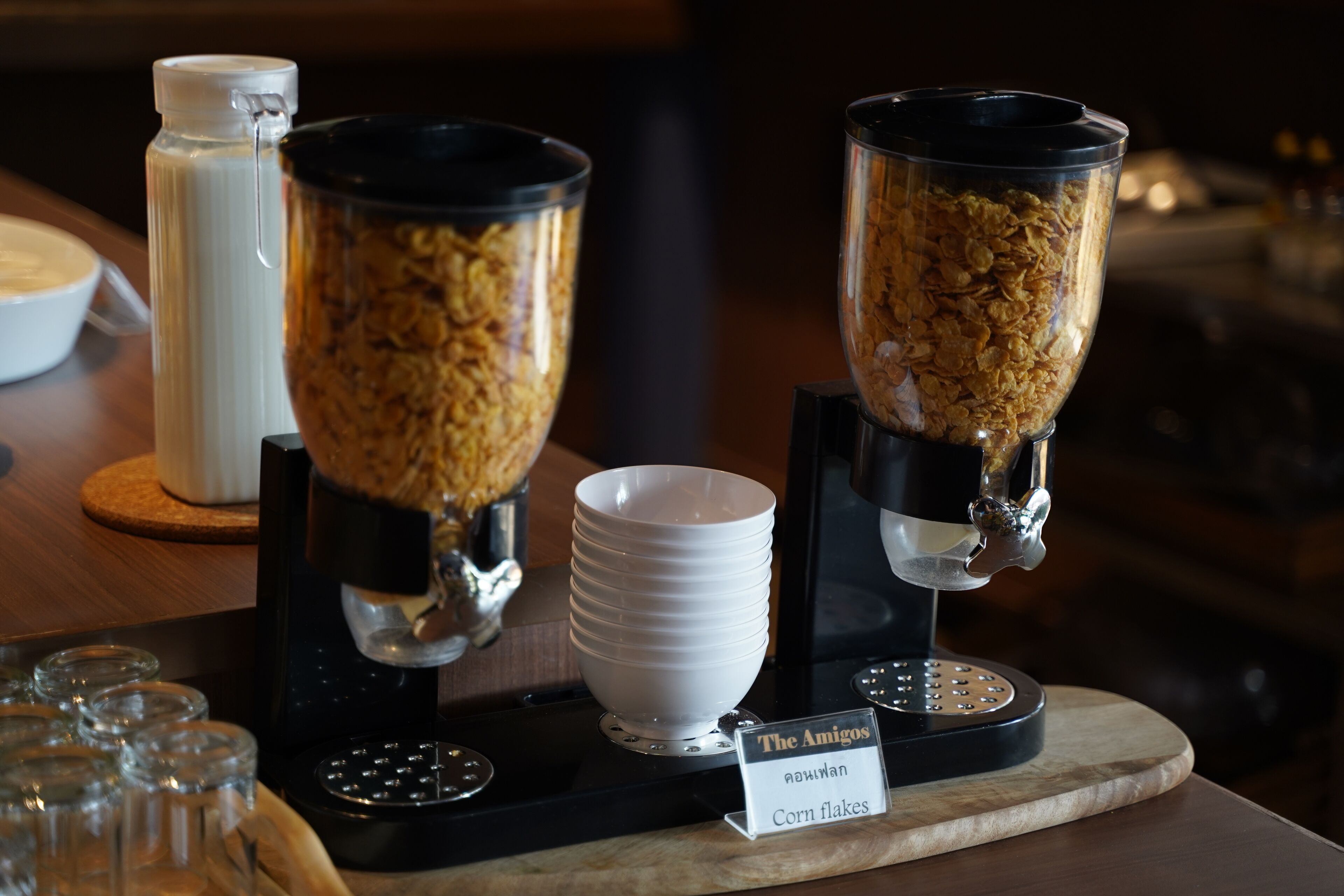 Estação de café da manhã com dois grandes recipientes de flocos de milho, um dispensador de leite e várias tigelas brancas.