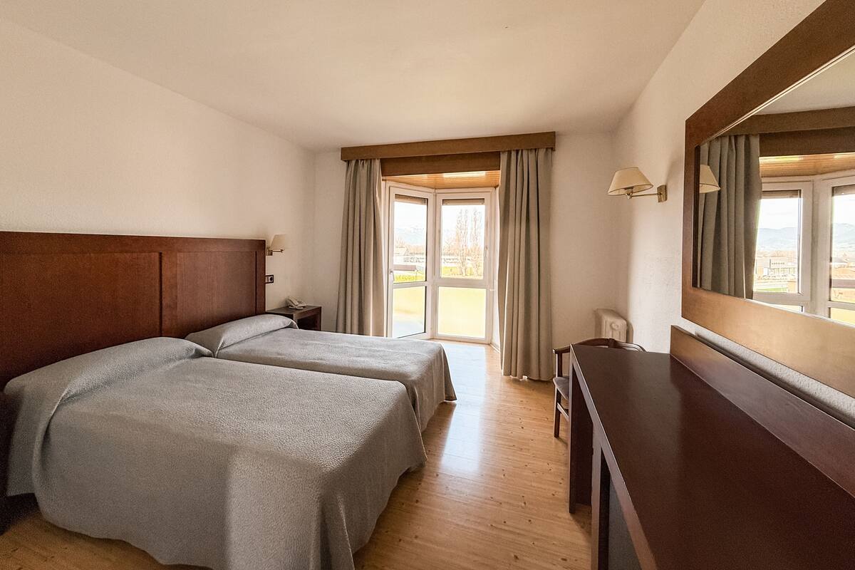 A hotel room with two beds, a desk, a mirror, and a window with curtains.