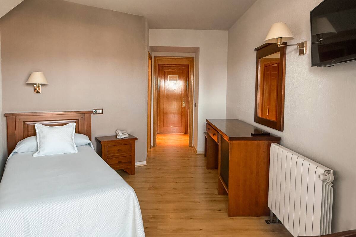 A hotel room with a bed, bedside table, lamp, dresser, mirror, and a TV mounted on the wall.