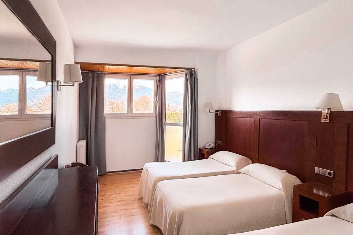 A hotel room with two beds, a wooden headboard, a desk, and a window with a view of mountains.