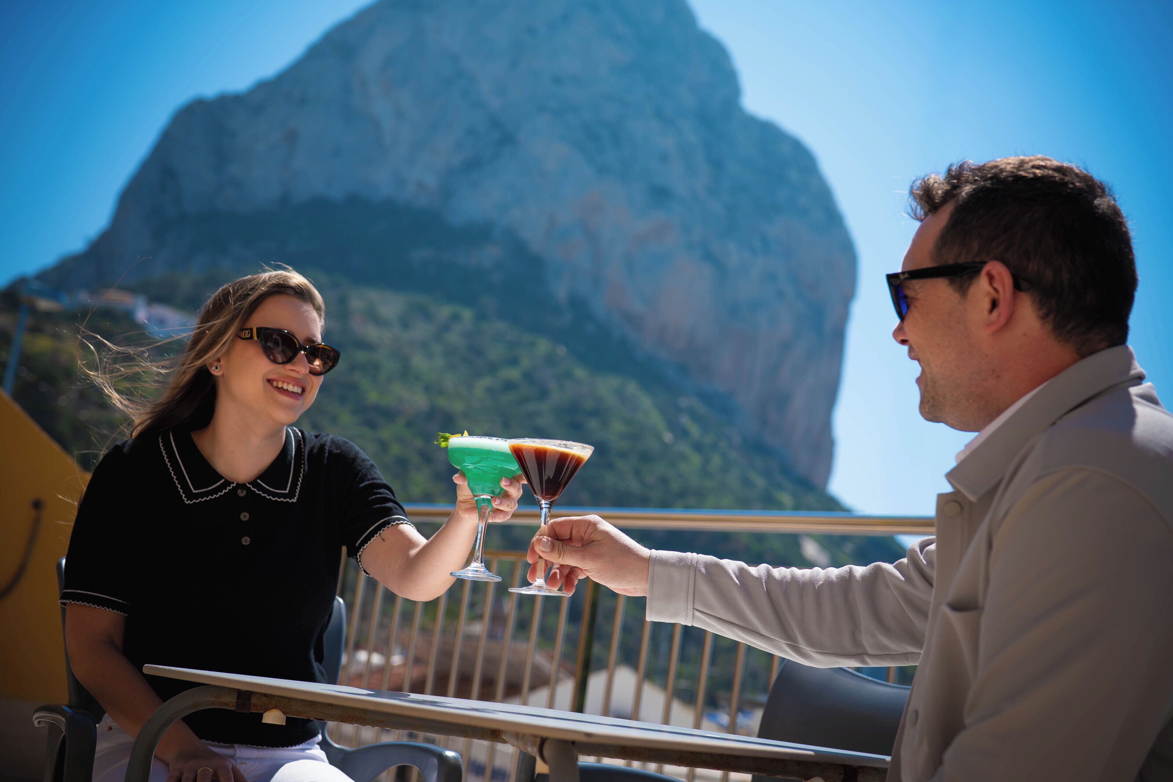 Una persona sosteniendo una copa de cóctel, otra persona ofreciendo un brindis, y un paisaje montañoso al fondo.