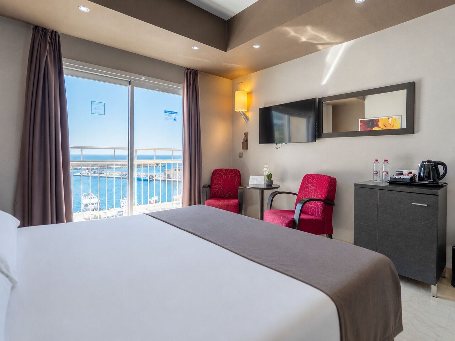 A hotel room with a bed, two red chairs, a small table with a lamp, a TV, and a view of the sea.