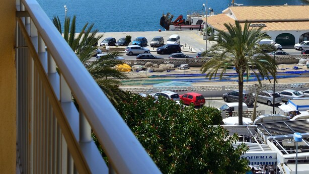 Vista costera desde un balcón con vista a un puerto deportivo con coches aparcados, un faro y un edificio con un cartel.
