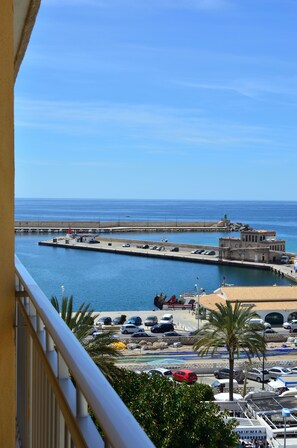 Vista costera desde un balcón con vista a un puerto deportivo con coches aparcados, un faro y un edificio con un cartel.