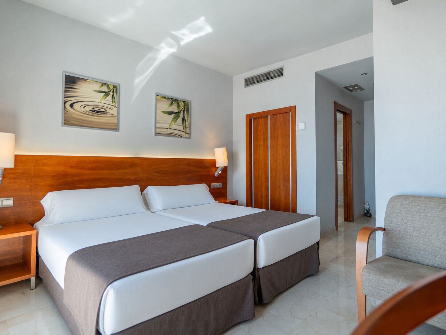 A hotel room with a double bed, two bedside tables, a wooden headboard, and two wall-mounted paintings.