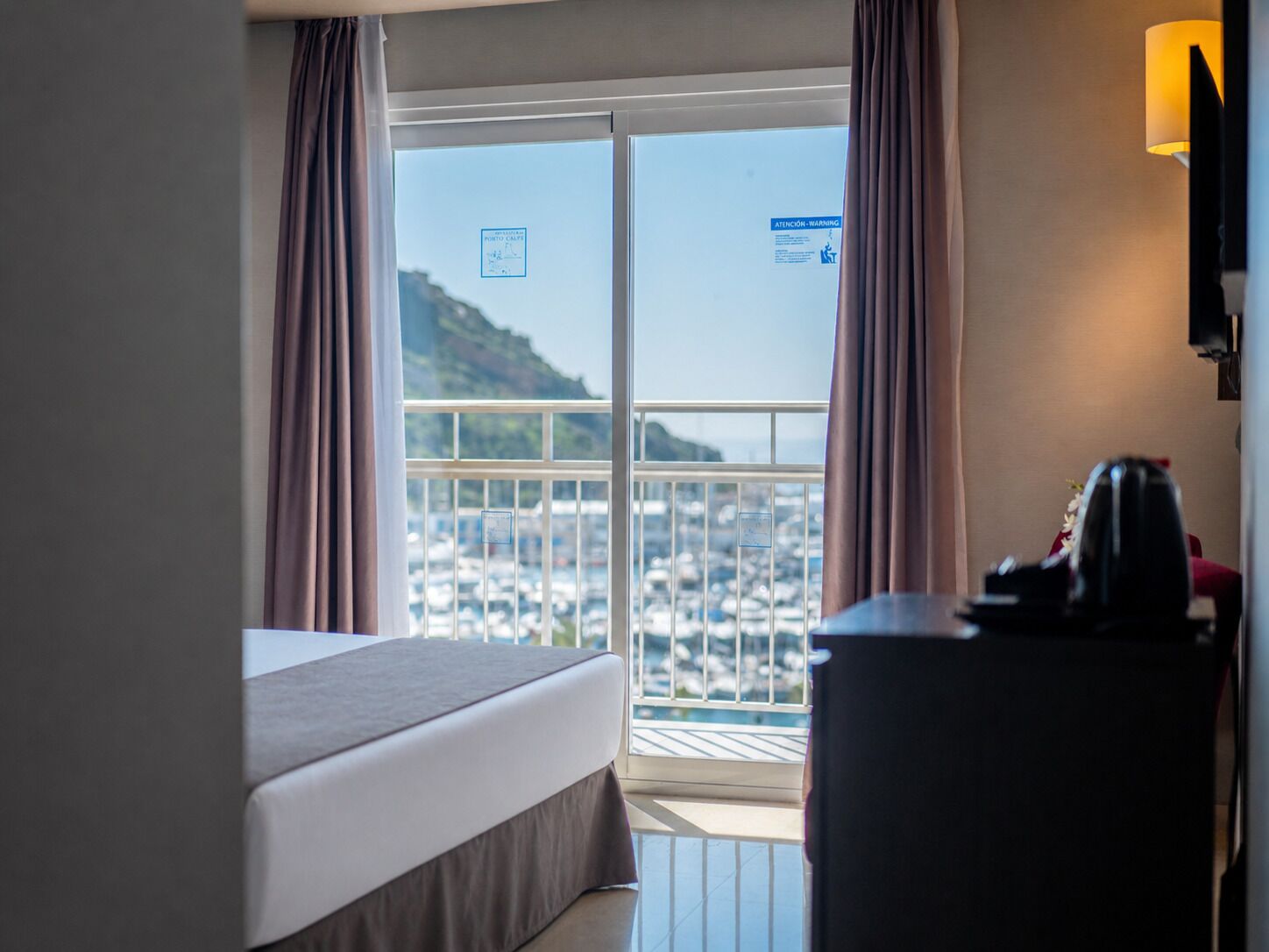 Een hotelkamer met een bed, een bureau en uitzicht op een jachthaven en heuvels.