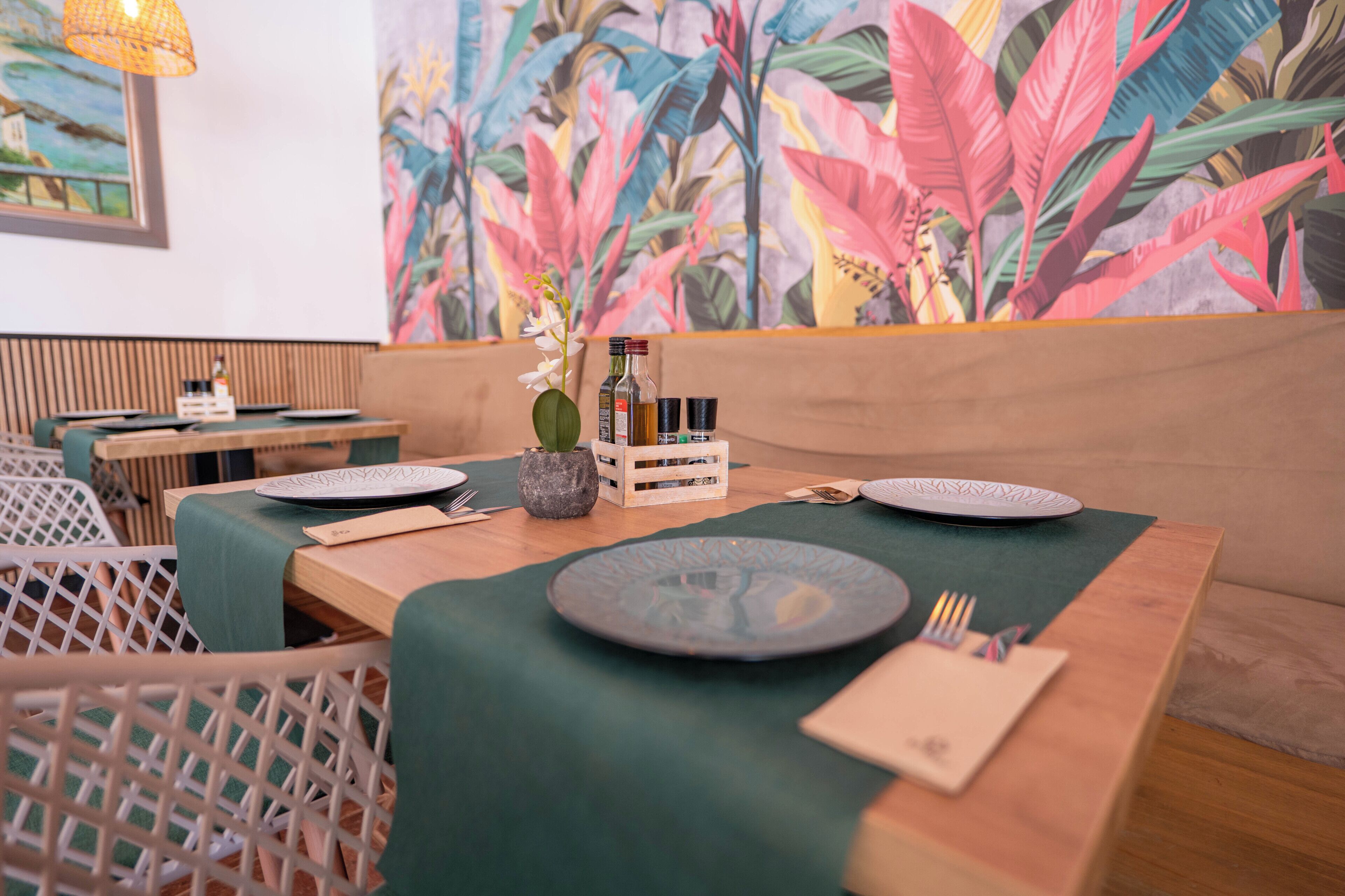 A dining area with a wooden table set for two, a green tablecloth, and a tropical plant mural in the background.