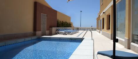 A swimming pool with lounge chairs and a large umbrella, surrounded by a building with a beige facade and a clear blue sky.