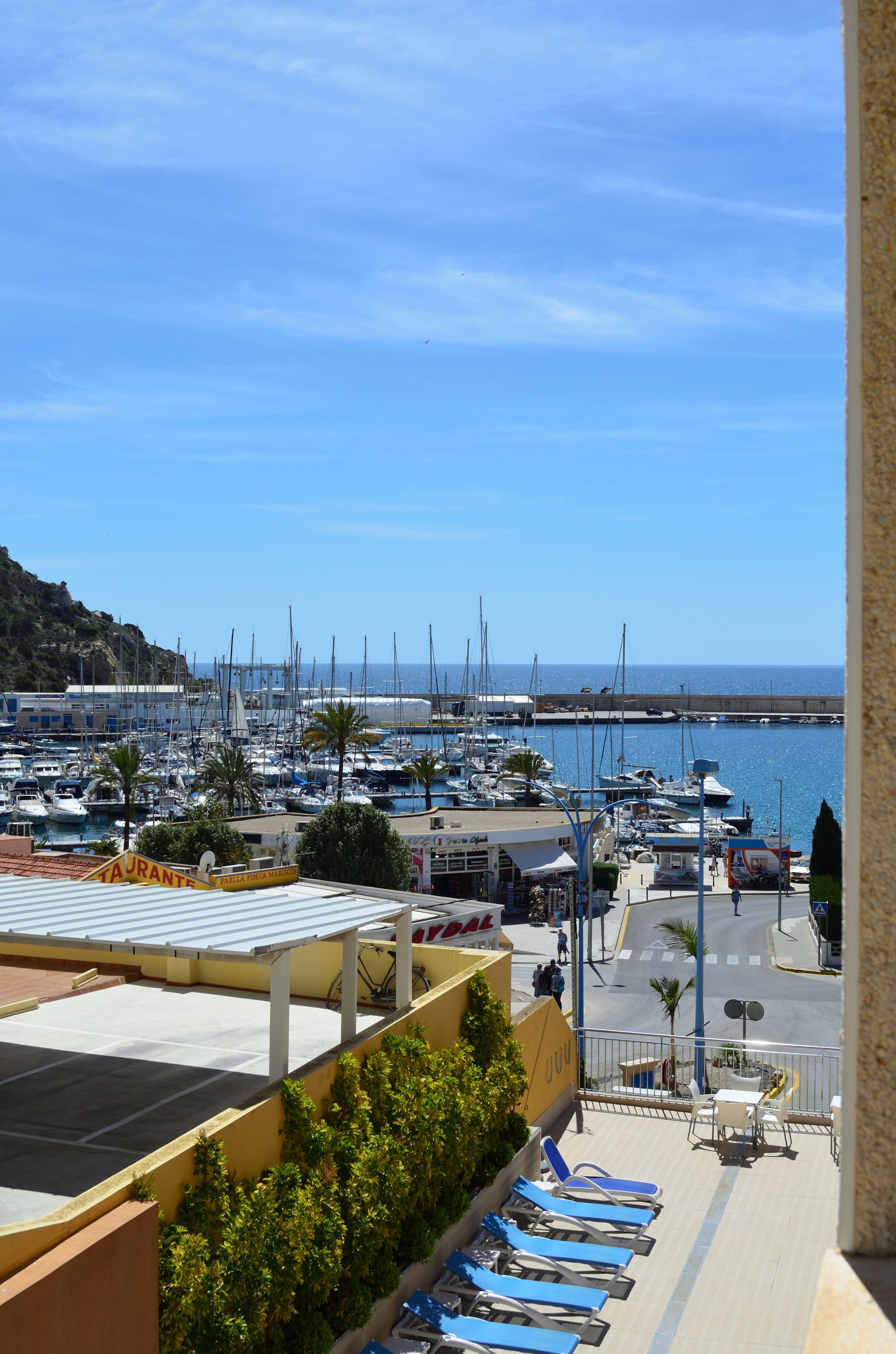 A marina with boats docked, a café with outdoor seating, and a view of the sea.