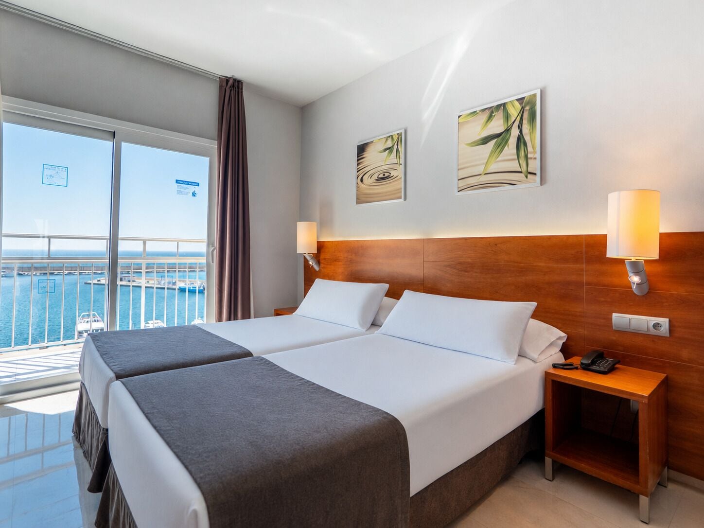 Ein Hotelzimmer mit zwei Betten, einem Holz-Kopfteil, einem Nachttisch mit Telefon und Meerblick durch große Fenster.