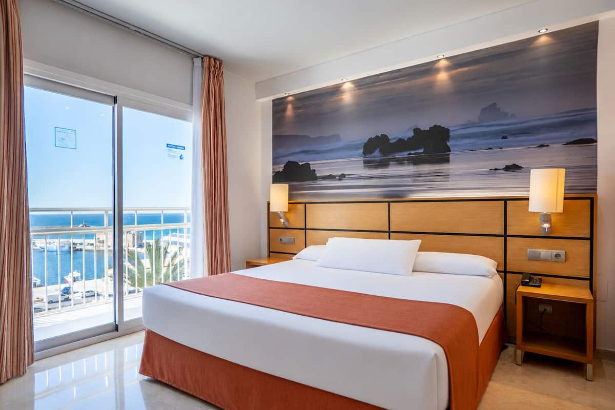 A hotel room with a large bed, a view of the sea, and a painting of a beach scene.