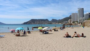 Una playa con gente descansando bajo sombrillas, un cielo despejado y montañas al fondo.