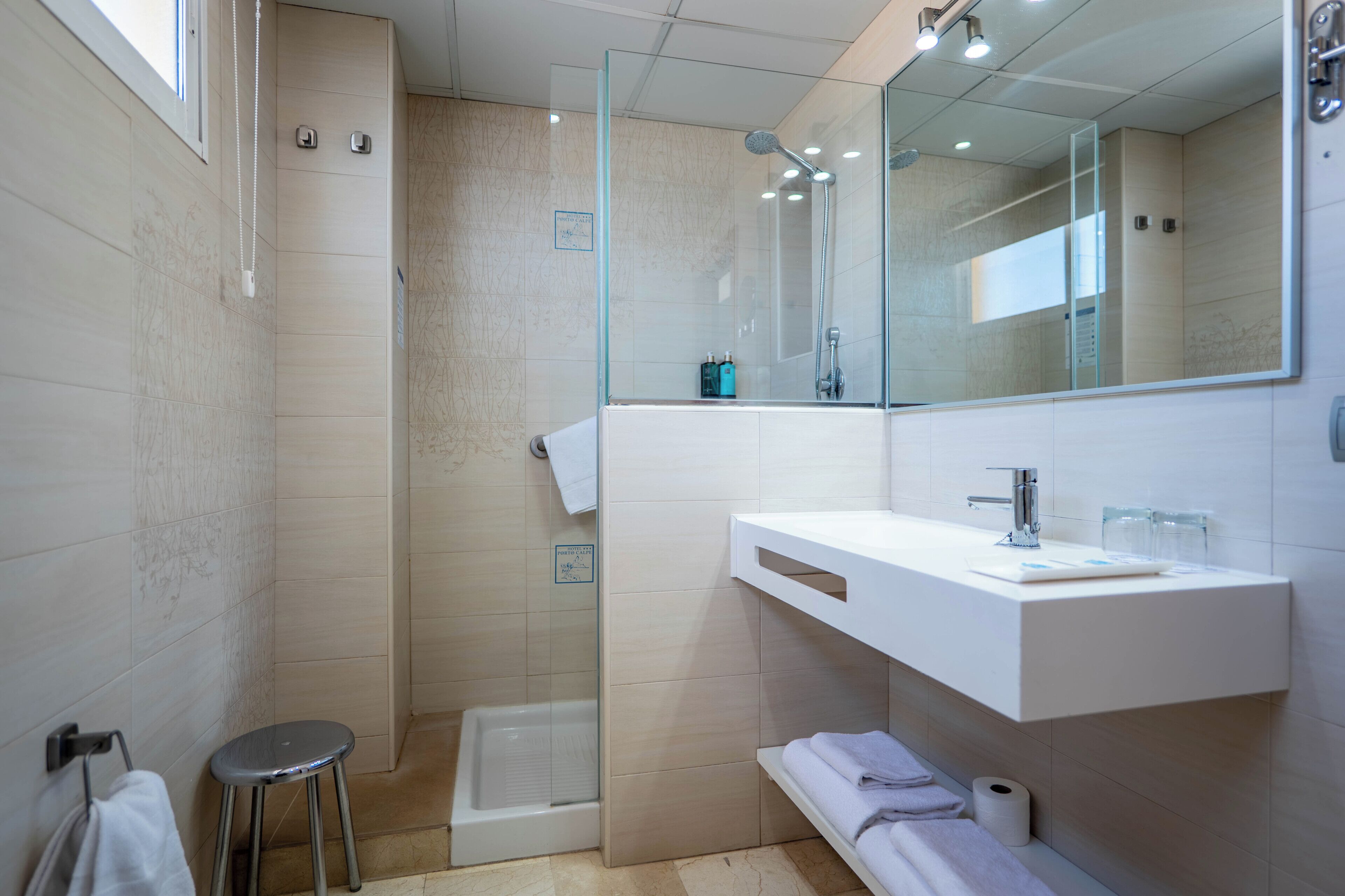 A modern bathroom with a glass-enclosed shower, a white vanity with a mirror, and a small stool.