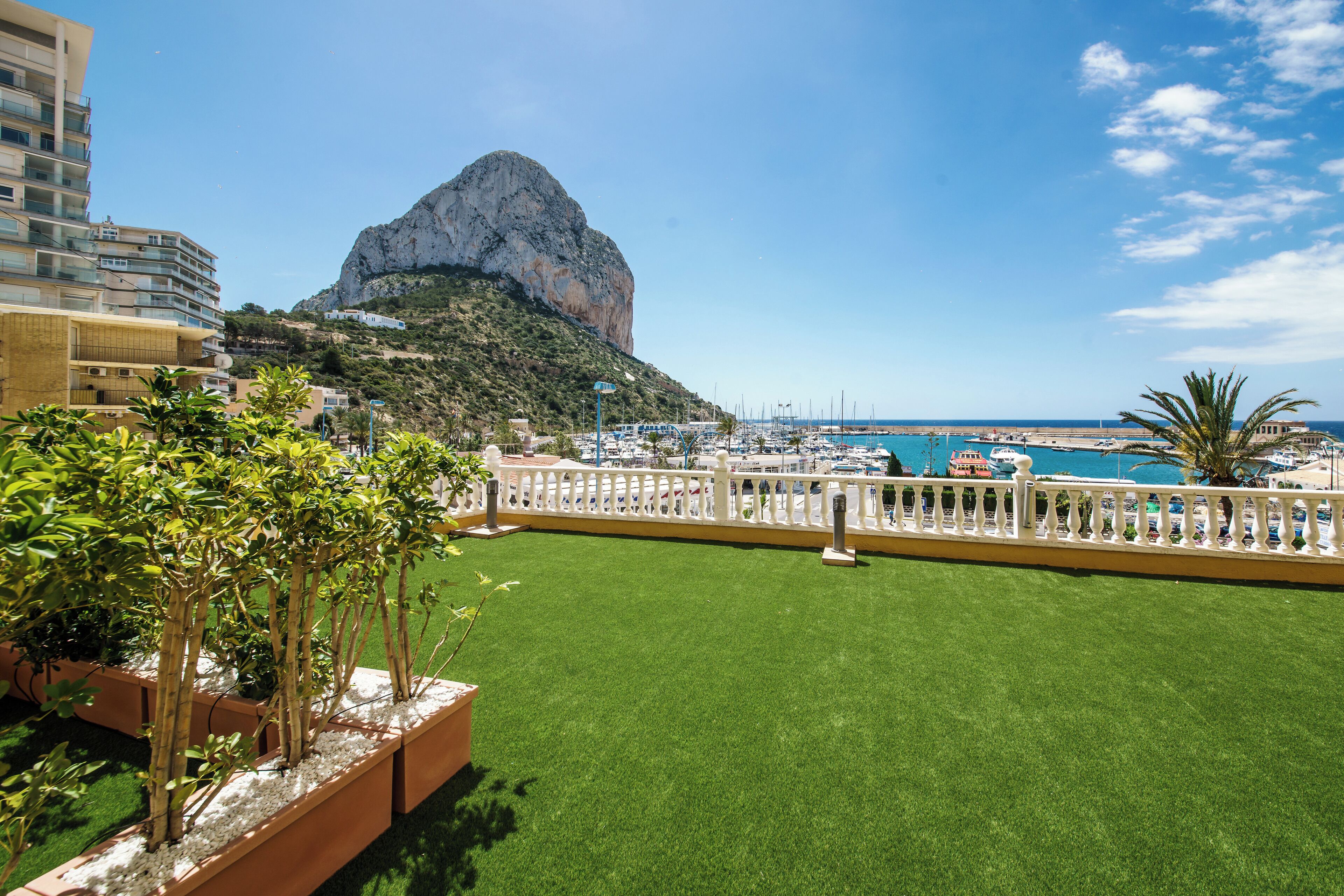 Una terraza en la azotea con césped artificial, barandilla blanca y vistas a un puerto deportivo con barcos y una montaña al fondo.